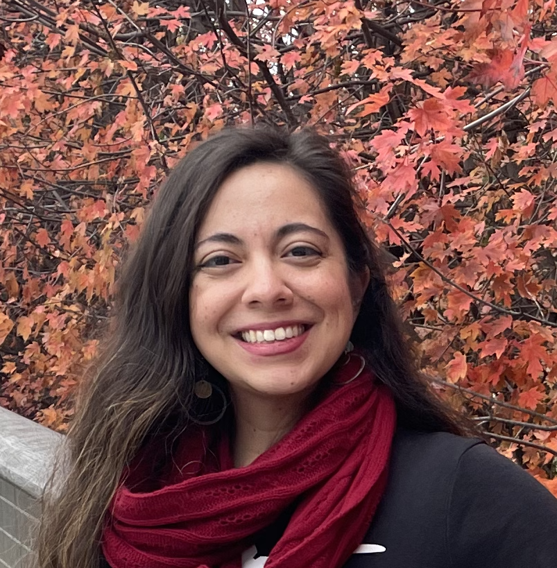 Corrine Rojas wearing a red scarf while smiling in front of a red-leafed bush.