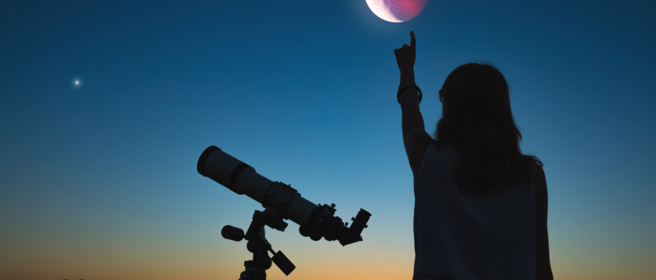 
		Silhouette of a person standing next to a telescope during twilight, pointing at the sky. The sky is transitioning from sunset to night, displaying a gradient of colors from deep orange near the horizon to dark blue higher up. A large, detailed moon with a reddish hue is prominently visible in the sky. A single bright star or planet can be seen in the distance. The scene conveys a sense of wonder and exploration.		