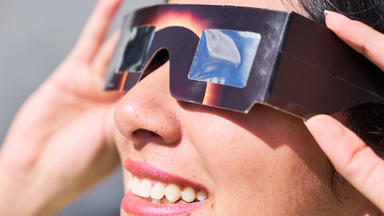 
		Close-up of a person smiling while wearing protective eclipse glasses. The glasses are designed with a dark frame and reflective lenses to safely view a solar eclipse. The person's hands are adjusting the glasses, and their face is illuminated by natural light. The detailed reflection of the sky can be seen on the lenses. The image captures the excitement and wonder of observing a solar eclipse.		